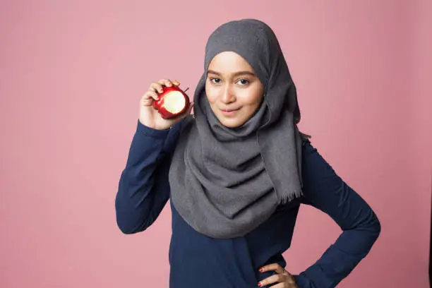 A young beautiful hijab sportsgirl is holding an apple looking at camera isolated on a pink background