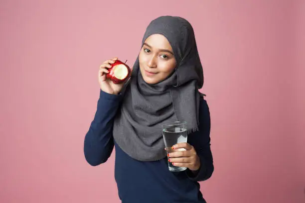 A young beautiful hijab sportsgirl is biting an apple with plain water looking at camera isolated with pink background