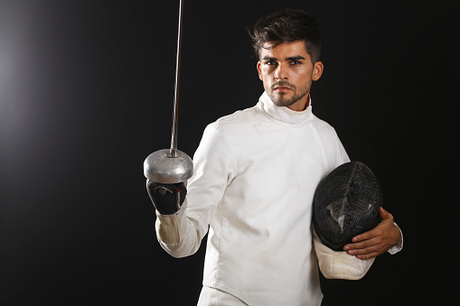 Young man in fencing equipment - uniform, face mask and foil sword on black background. About 20 years old, Latin man.