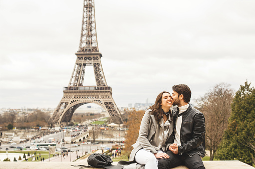 Young couple sitting on surrounding wall with a view on Eiffel tower, the symbol of France. The man kisses his girlfriend on the cheek.