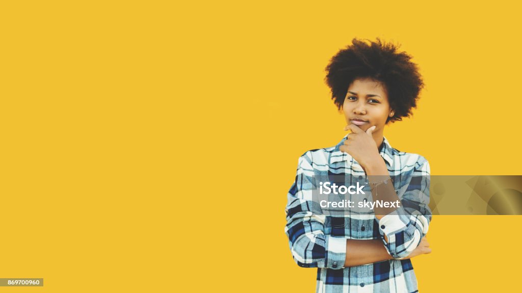 Pensive brazilian girl on isolated yellow background Young black female teenager in plaid shirt is making pensive emotional face while standing in front of isolated solid yellow background with copy space place for logo, text or your advertising message People Stock Photo