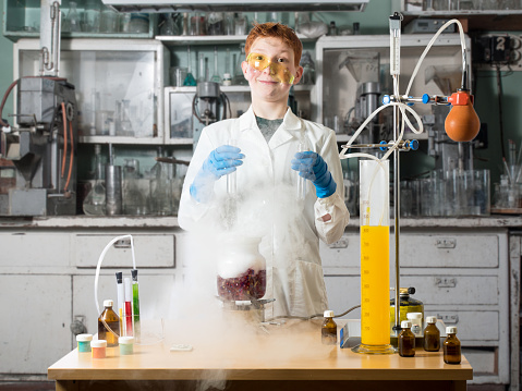 Schoolboy with move out on his nose glasses stands in a white robe beside chemical table and holds in hands of the flask on a background of chemical laboratory with table and chemical equipment