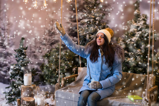 istock Young attractive woman in blue down jacket greeting somebody while sitting on a swing in a snow-covered park 869709746