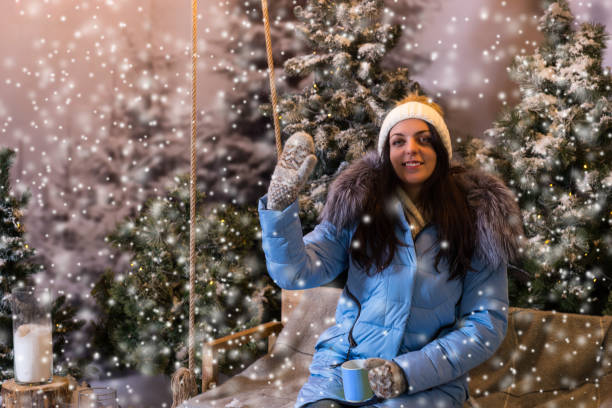 istock Young woman in blue down jacket greeting while sitting on a swing in a snow-covered park 869710144