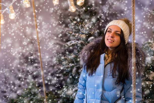 Cute young woman in blue down jacket standing in a snow-covered park with spruce trees while snowing