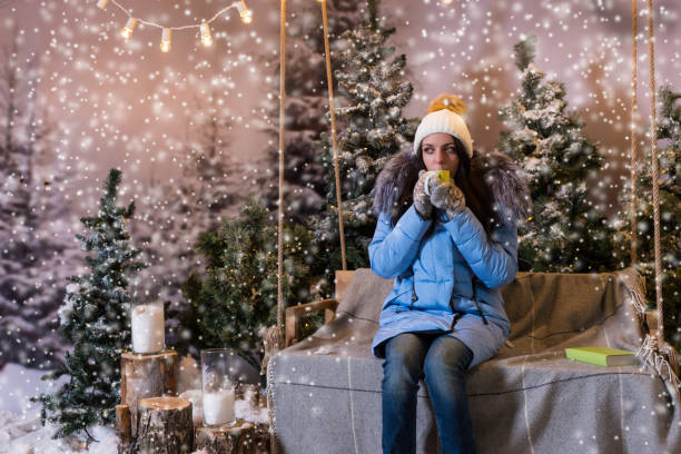 Woman in blue down jacket drinking hot drink while sitting on a swing under the flashlights in a snow-covered park