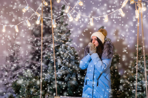 istock Female in blue down jacket warming hands in mittens under the flashlights in a snow-covered park 869714346
