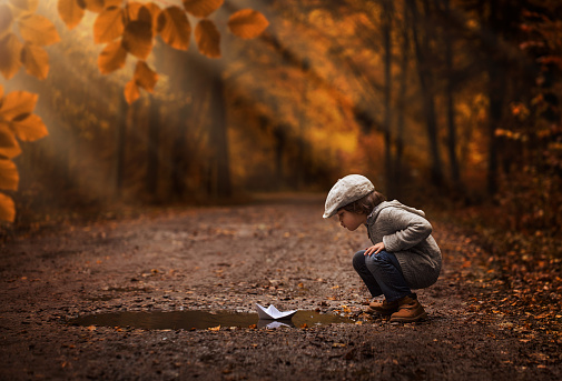 Little boy playing with the paper boat in the autumn forest puddle