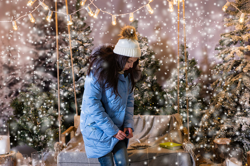 Cute woman buttoned up her blue down jacket and standing near swing with a blanket under the flashlights in a snow-covered park, wearing warm woolen knitted scarf and hat while snowing, concept about Christmas and New Year holiday