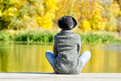 Girl in a hat and coat sitting on the dock. Autumn, sunny. Back view