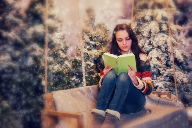 istock Young woman reading a book while sitting on a swing with her legs up on it in a snow-covered park 869726174