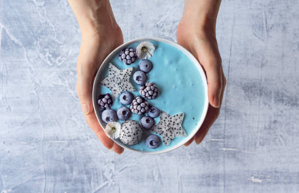 Woman's Hands Holding Blue Spirulina Yogurt Smoothie Bowl with Berries and Dragon Fruit An anonymous woman's hands holding a blue smoothie bowl topped with frozen berries and dragon fruit. The smoothie bowl is made with yogurt, blue spirulina powder, and fresh fruit (blueberries and blackberries). spirulina stock pictures, royalty-free photos & images