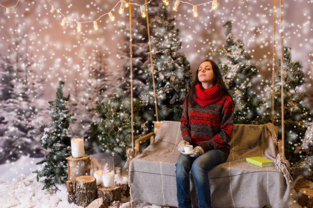 istock Young dreaming female sitting on a swing with a blanket under the flashlights and holding a cup of coffee in a snow-covered park 869767072
