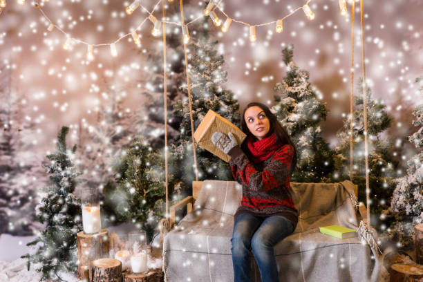 istock Woman sitting on a swing with a blanket under the flashlights and shaking a box with a present in a snow-covered park 869771920