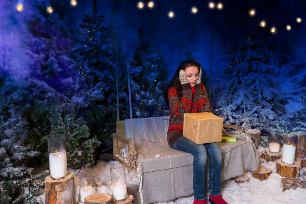 istock Surprised woman sitting on a swing with a blanket under the flashlights and holding a box with a present in a snow-covered park in the evening 869772502