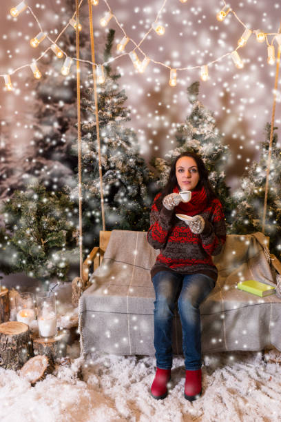 istock Young female sitting on a swing with a blanket under the flashlights and holding a cup of coffee in a snow-covered park 869773936