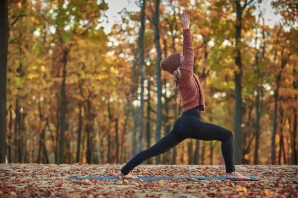 Beautiful young woman practices yoga asana Virabhadrasana 1 - warrior pose on the wooden deck in the autumn park