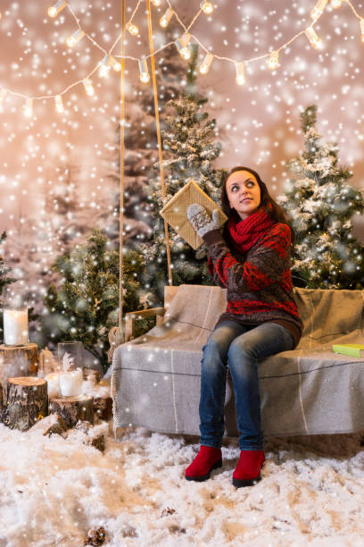istock Cute girl sitting on a swing with a blanket under the flashlights and shaking a box with a present in a snow-covered park 869779950