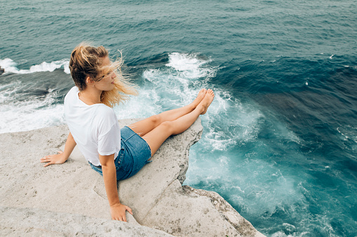 A young woman sits on the edge of a cliff above the sea at Bondi Beach.