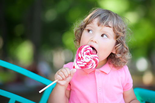 Beautiful little girl holding a big heart shaped lollipop, walking in the park, summer day.