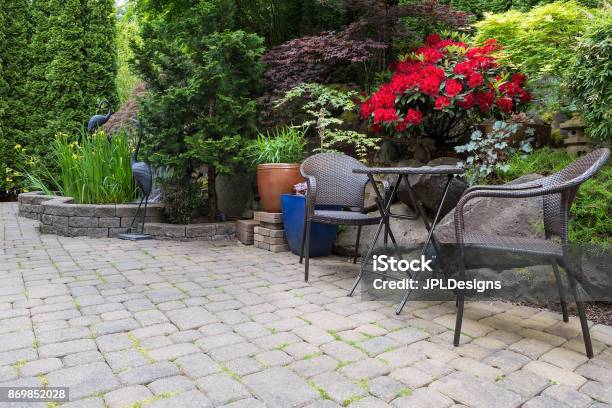 Garden Backyard With Lush Plants Landscaping Pond Water Fountain And Stone Paver Patio Hardscape With Wicker Bistro Furniture Chair And Table Set Stock Photo - Download Image Now