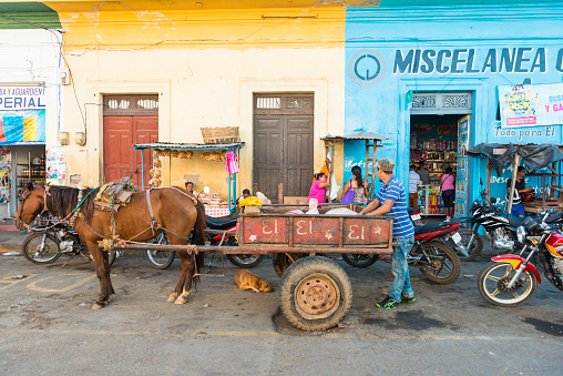 A colorful streetview in old part of Granada in the morning. In front a van drawn with horse, busy sidewalk in front of a grocery shop. Nicaragua.