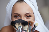 Young beautiful smiling woman with towel on her head holding make-up brushes in her hands.