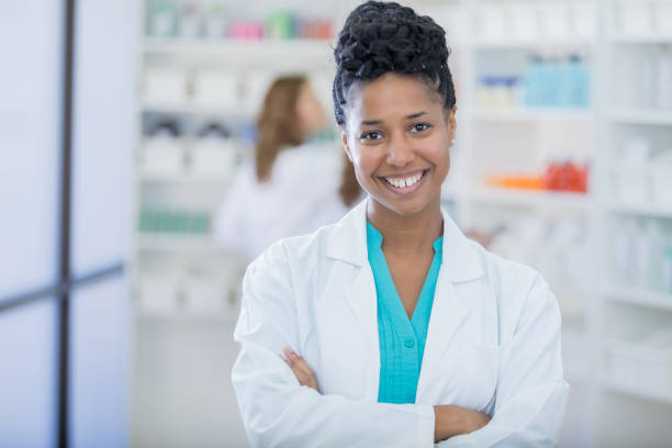 Portrait of beautiful female pharmacist Confident mid adult African American pharmacist stands in her pharmacy with her arms crossed. She is wearing a lab coat. A pharmacist is working in the background. pharmacy tech stock pictures, royalty-free photos & images