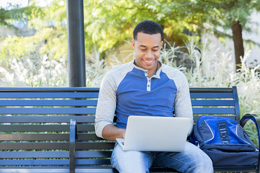A cheerful young male student sits on a metal bench outside alone on his college campus.  A backpack sits beside him as he looks down and types on his laptop.