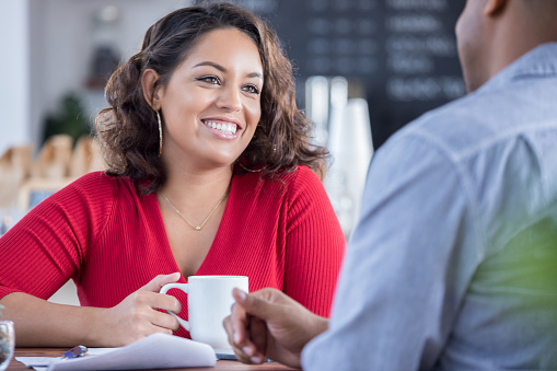 Beautiful young woman and an unrecognizable man are on a date in a local coffee shop. The woman is smiling while talking with the man. She is drinking a cup of coffee.