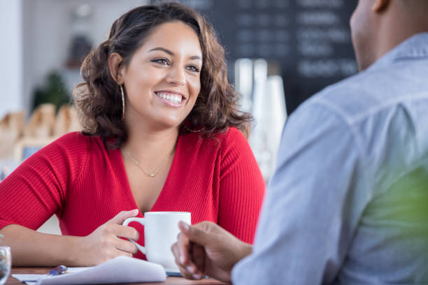 Young couple on a date in a local coffee shop Beautiful young woman and an unrecognizable man are on a date in a local coffee shop. The woman is smiling while talking with the man. She is drinking a cup of coffee. blind date stock pictures, royalty-free photos & images