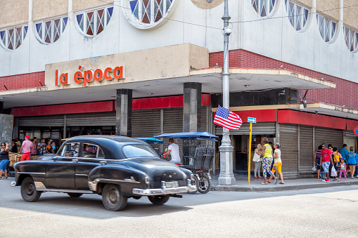 Pedestrians, shopppers, tourists walking down street on a sunny day in Havana Vieja, Cuba. Black vintage American car is visible in the foreground.\n\n