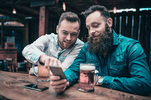 Two Smiling Bearded Hipster Friends Drinking Beer and Using Mobile Phone
