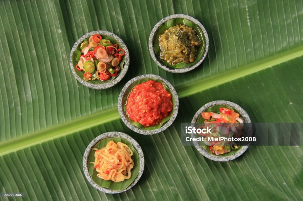 Five Indonesian Chili Relishes in Mini Stoneware Mortars Five popular relishes from Indonesia, plated in saucers of mini stoneware mortars. The saucers are arranged on a large banana leaf. The relishes are (in clockwise direction starting from top right) Sambal Lado Mudo / Sambal Cabe Ijo (Green Chili Paste) from West Sumatra; Sambal Dabu-Dabu (Fresh Chili and Tomato Salsa) from Sulawesi; Sambal Mangga (Unripen Mango Relish) from West Java; Sambal Matah (Raw Chili and Lemongrass Salsa) from Bali and in the center is Sambal Tomat (Tomato Chili Paste) popular across Java Island and Bali. Banana Leaf Stock Photo