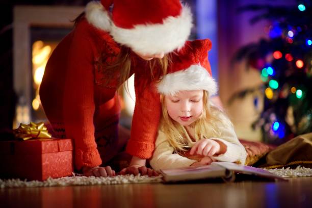 istock Two cute little sisters reading a story book together under a Christmas tree 869943662