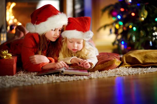 istock Two cute little sisters reading a story book together under a Christmas tree 869943694
