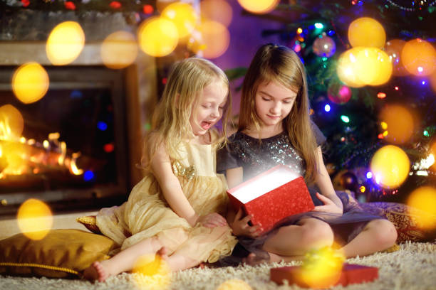istock Happy little sisters opening magical Christmas gift by a fireplace 869950156