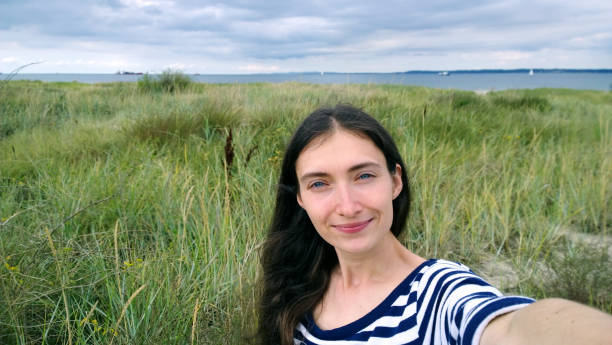 Brunette girl in the striped shirt taking a selfie on the beach. Sandy beach with dunes. Wind cloudy cool weather stock photo