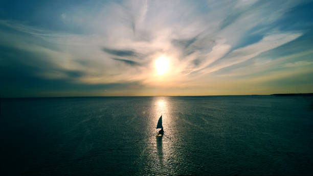 Little white boat floating on the water towards the horizon in the rays of the setting sun. Beautiful clouds with yellow highlights. Aerial view stock photo