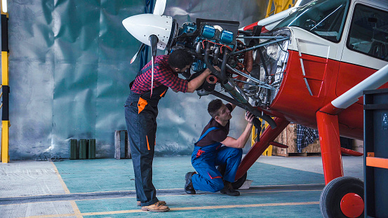 Two mechanics working on a small aircraft in a hangar with the cowling off the engine as they perform a service or repair