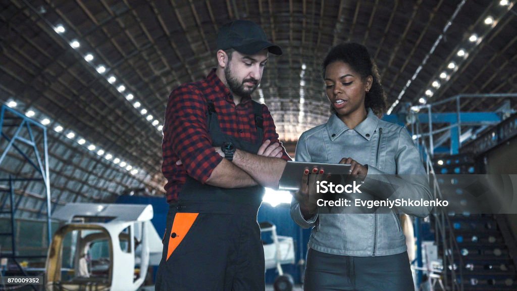 Man discussing in aircraft hangar with woman Man discussing over digital tablet in aircraft hangar with woman Air Vehicle Stock Photo