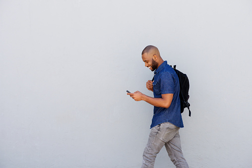 Side portrait of a happy young guy with bag walking outdoors and sending text message by mobile phone