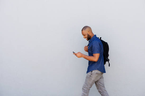 happy young guy with bag walking with mobile phone Side portrait of a happy young guy with bag walking outdoors and sending text message by mobile phone men hairstyle afro cool stock pictures, royalty-free photos & images