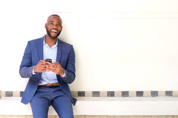 smiling african business man sitting against wall with mobile phone Portrait of smiling african business man sitting against wall with mobile phone businessman african descent on the phone business person stock pictures, royalty-free photos & images