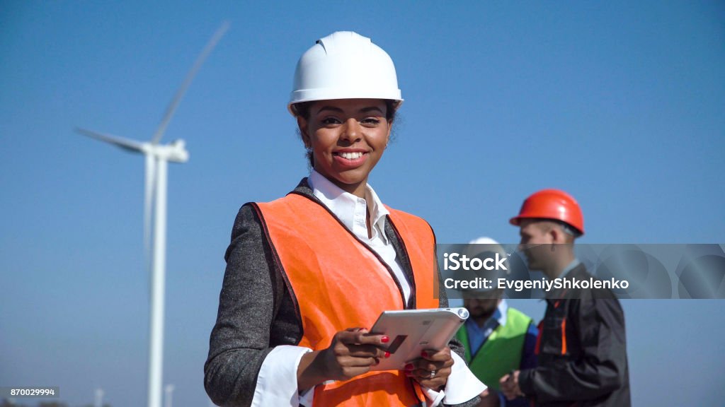 Woman with protective helmet against wind turbine Female engineer wearing hardhat standing with digital tablet against other engineers and wind turbine on sunny day and looking at camera Engineer Stock Photo