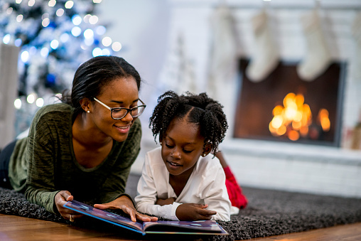 A mother and daughter of African descent are in indoors in their living room during Christmas time. They are wearing warm, comfortable clothes. They are laying on the carpet and reading a storybook together.