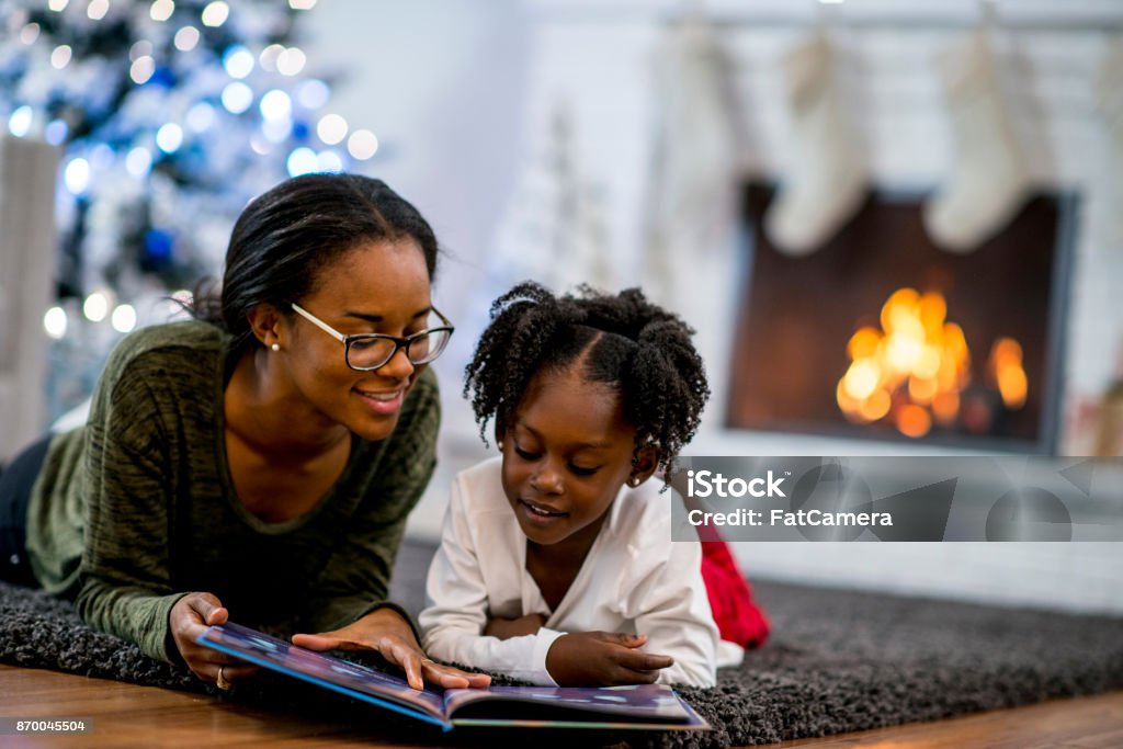 Storytime A mother and daughter of African descent are in indoors in their living room during Christmas time. They are wearing warm, comfortable clothes. They are laying on the carpet and reading a storybook together. Christmas Stock Photo