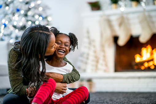 A mother and daughter of African descent are in indoors in their living room during Christmas time. They are wearing warm, comfortable clothes. The mother is hugging and kissing her daughter, and she is laughing.