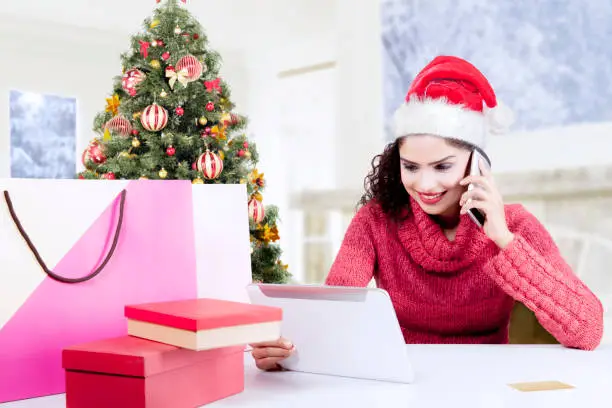 Happy woman wearing a Santa hat and buying Christmas presents online with a tablet computer while making a phone call at home