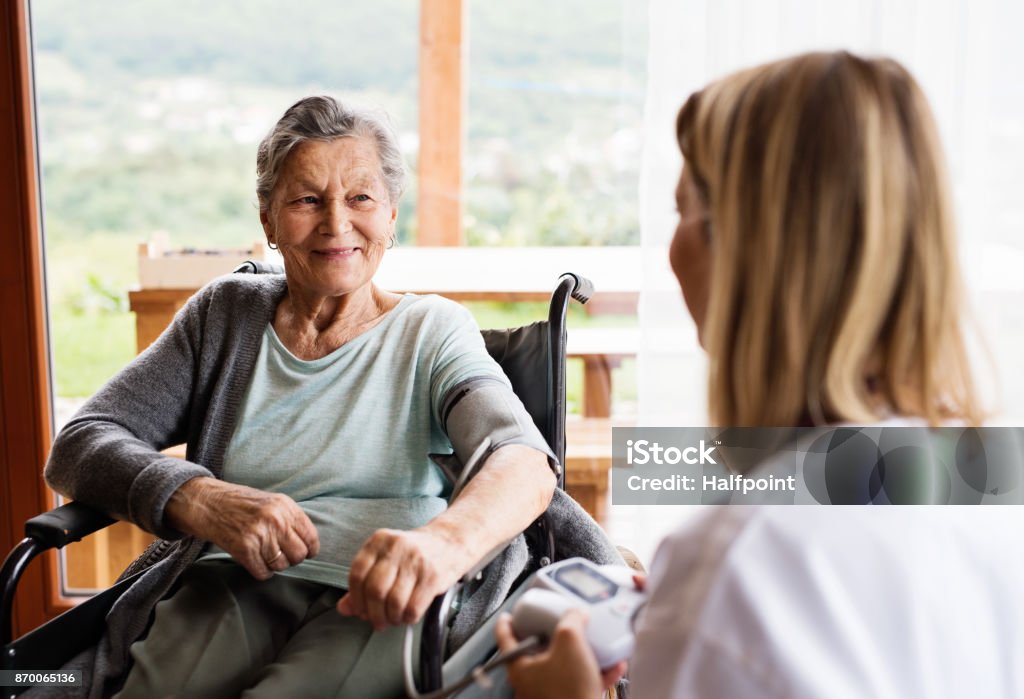 Health visitor and a senior woman during home visit. Health visitor and a senior woman during home visit. A nurse checking blood pressure of a woman in an wheelchair. Care Stock Photo